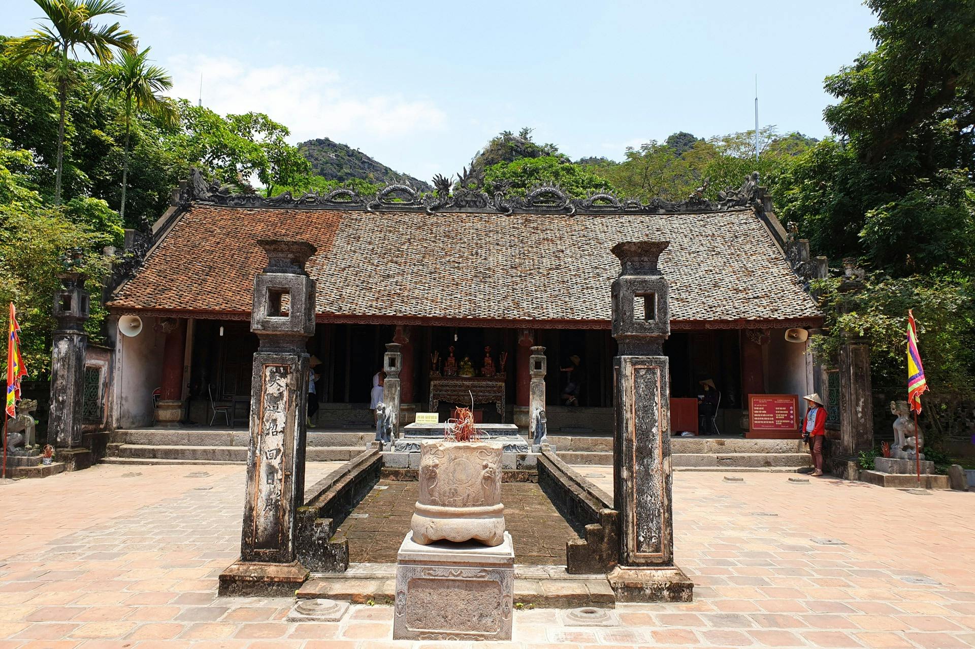 나무와 언덕을 배경으로 타일로 장식된 지붕, 석조 기둥, 향로, 안뜰의 인물이 있는 고대 사원.