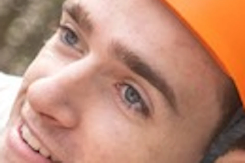 A person wearing a harness and orange helmet climbs a rope in a treetop adventure course surrounded by greenery.