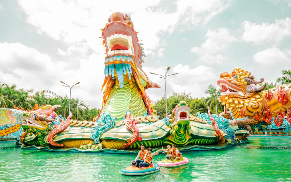 People on paddle boats in a colorful amusement park with giant dragon sculptures and a water setting under a cloudy sky.