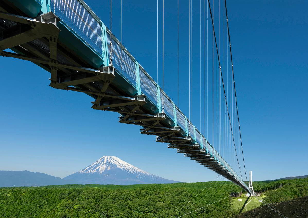 A suspension bridge with a blue structure spans over a lush green forest, with a snow-capped mountain in the background.