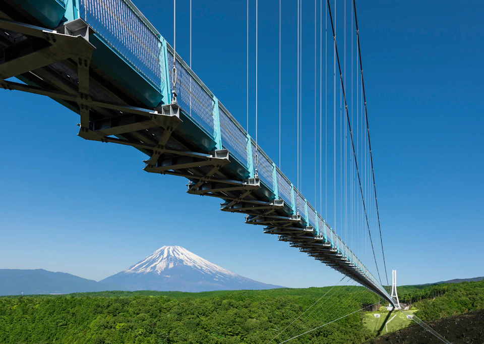 A suspension bridge over a green forest with a clear view of a snow-capped mountain in the distance under a blue sky.