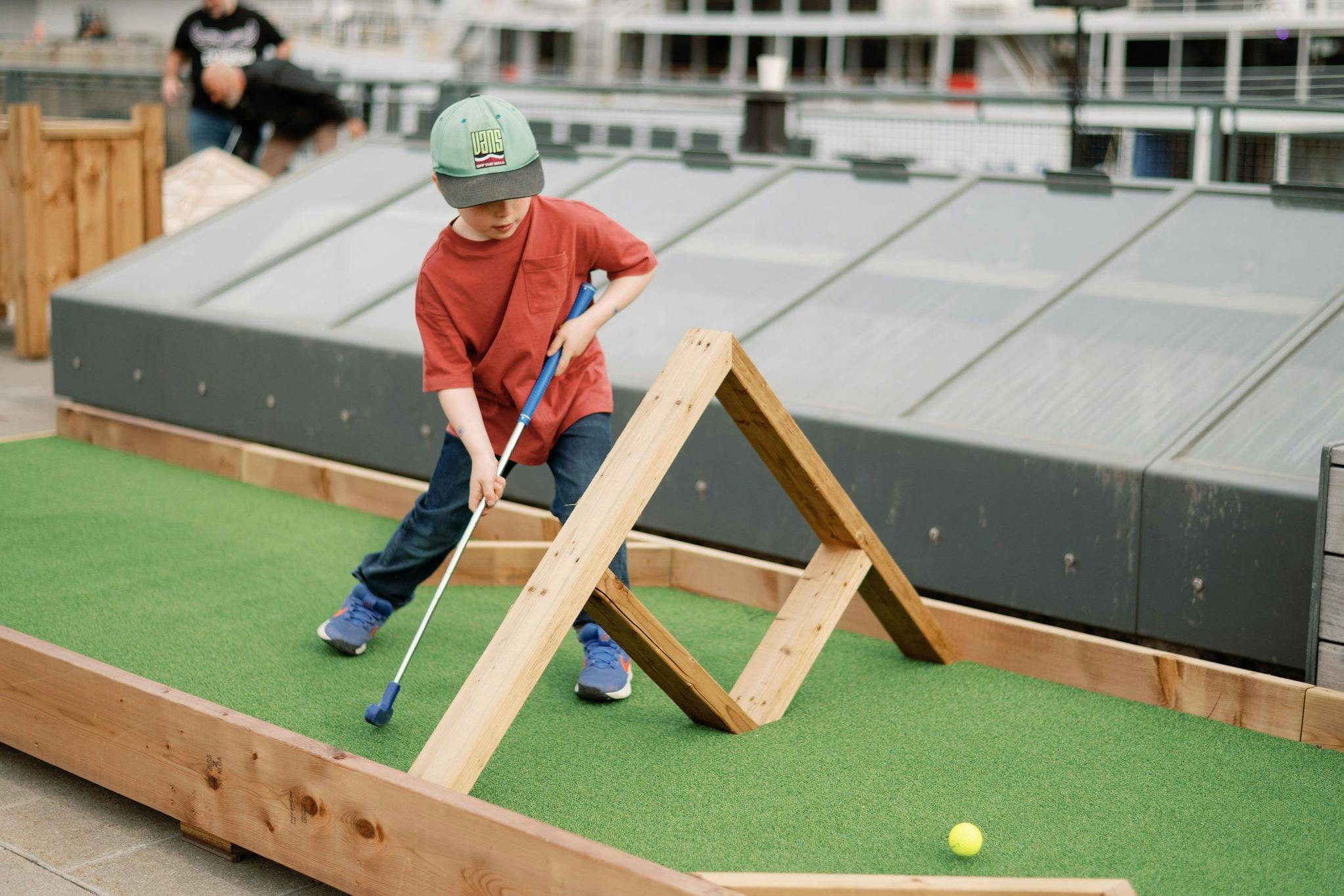 Uma criança a jogar mini-golfe num campo no telhado com obstáculos de madeira, com roupa informal e um boné.