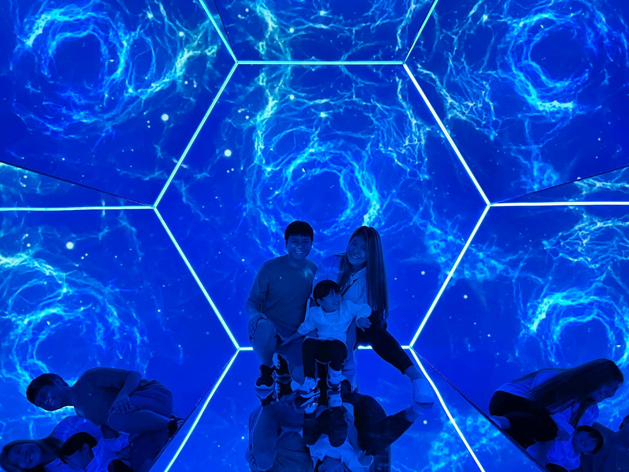 Two adults and a child pose inside a hexagonal room with glowing blue, cosmic-like patterns on the walls.