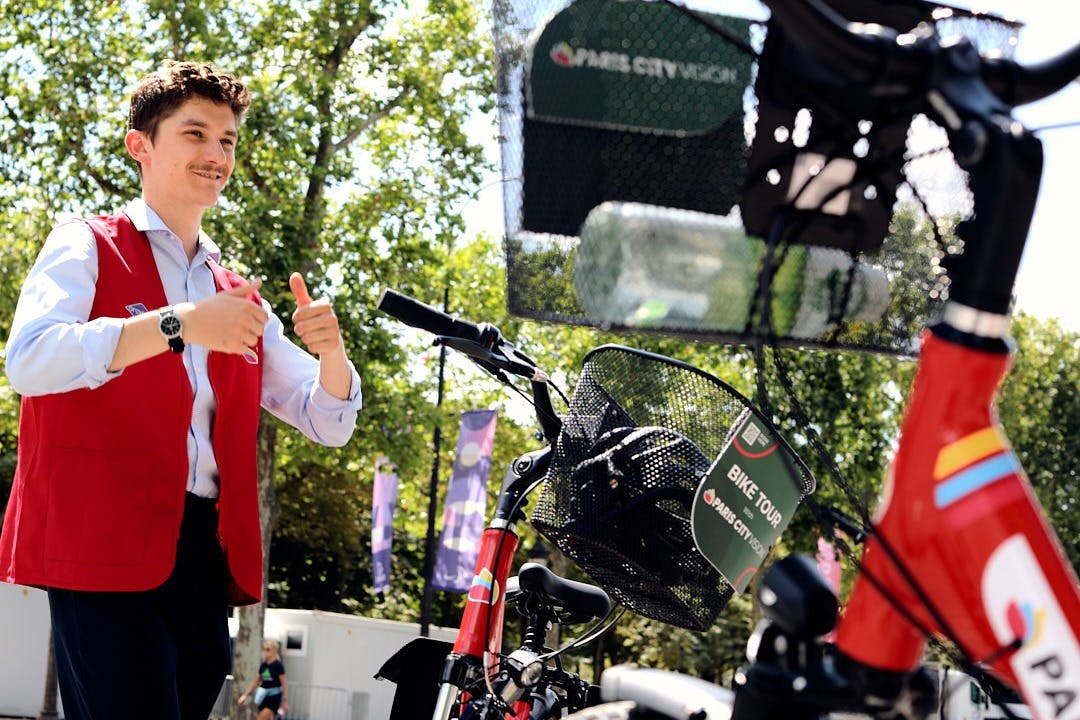 A person in a red vest giving thumbs up near a bicycle in an outdoor area with trees and banners in the background.