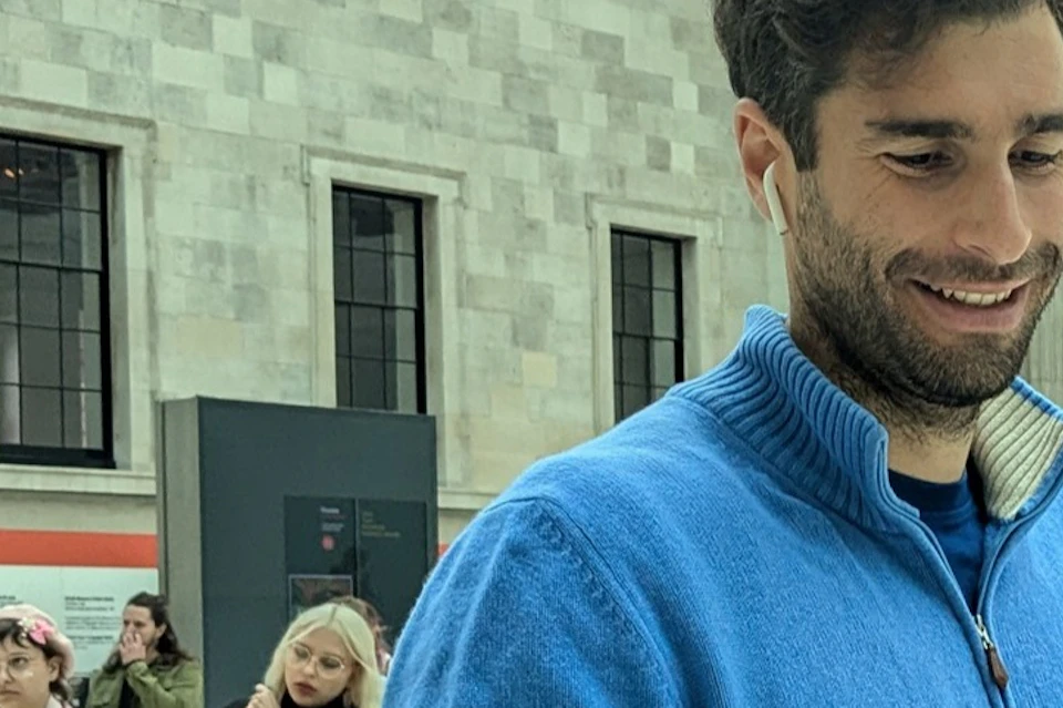 A man in a blue sweater looks at his phone in a large indoor space with architectural details and other people around.
