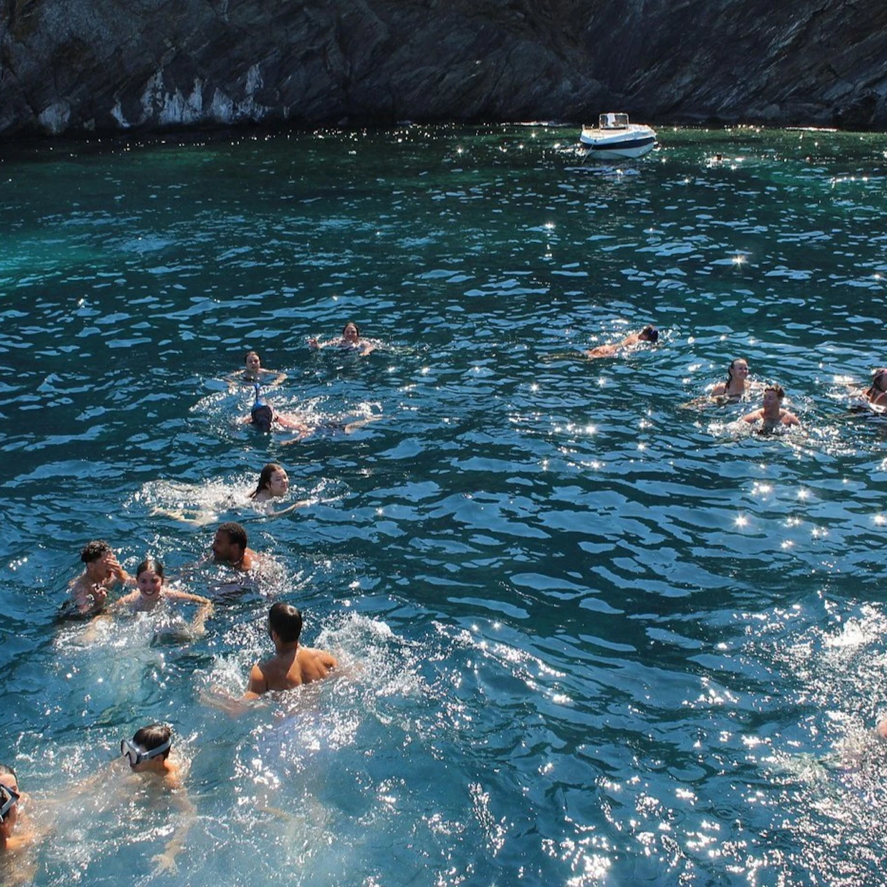 People swimming in a clear blue ocean near a rocky shoreline, with a small boat floating in the distance.