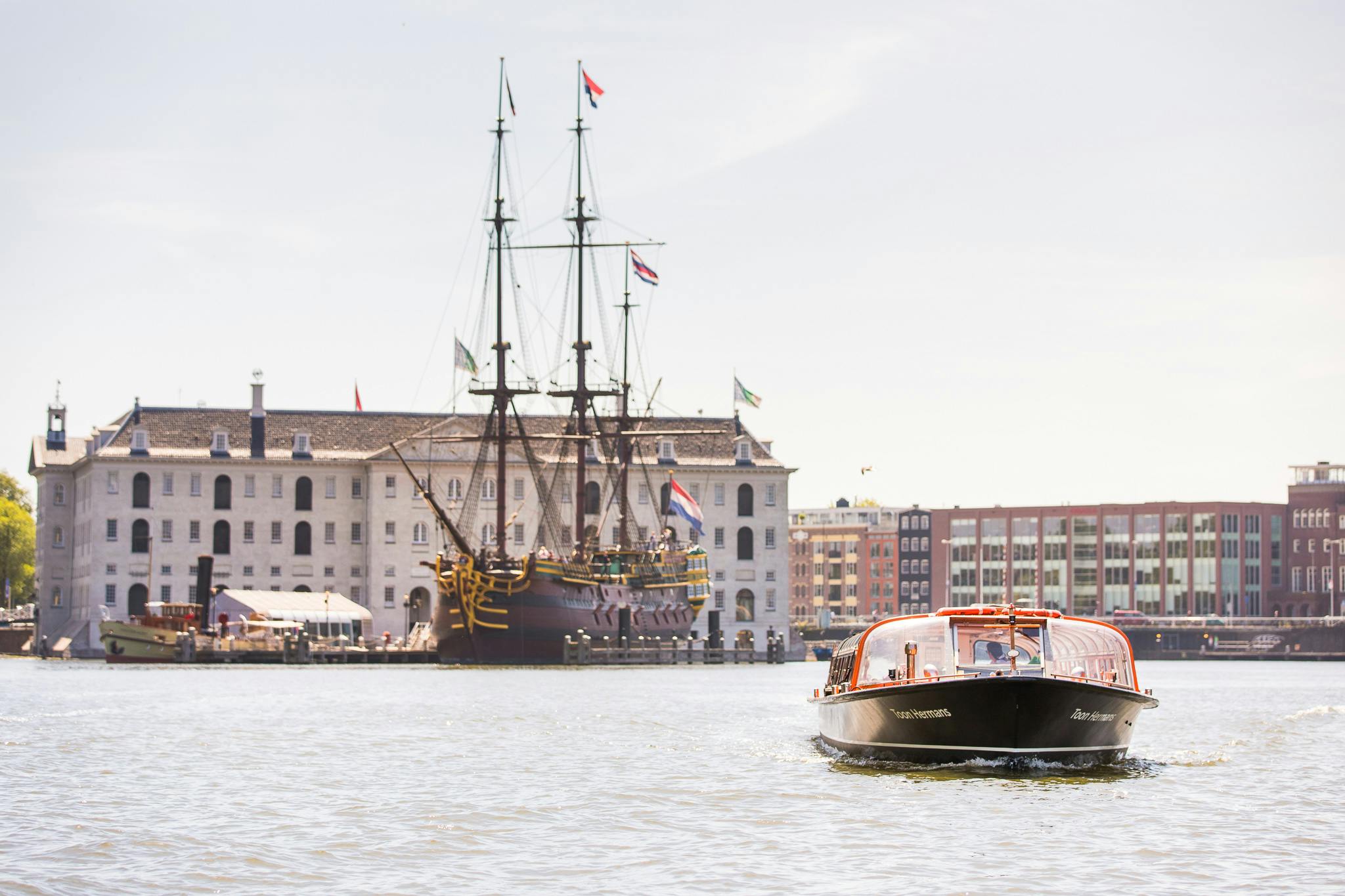 Ein Touristenboot auf einem Kanal fährt an einem angedockten historischen Schiff und einem großen Gebäude mit Flaggen im Hintergrund vorbei.
