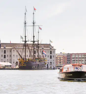 Een toeristenboot op een kanaal vaart langs een aangemeerd historisch schip en een groot gebouw met vlaggen op de achtergrond.