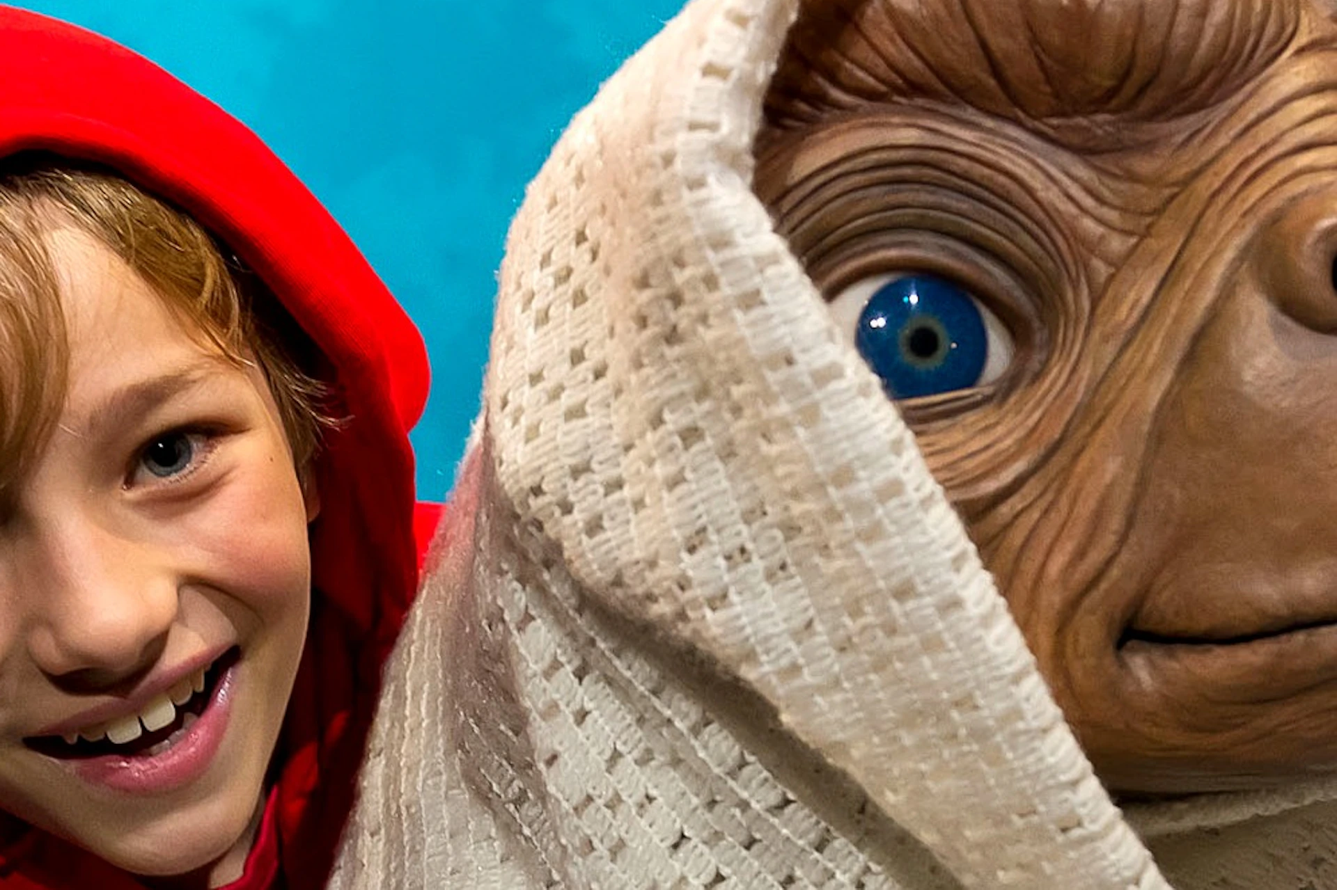 Smiling child in a red hoodie poses with a figure resembling an extraterrestrial wrapped in a white blanket against a blue background.