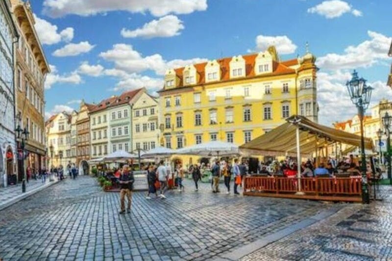 青く曇った空の下、カラフルな歴史的建造物に囲まれた屋外カフェで、人々が歩いたり食事をしたりする石畳の広場。