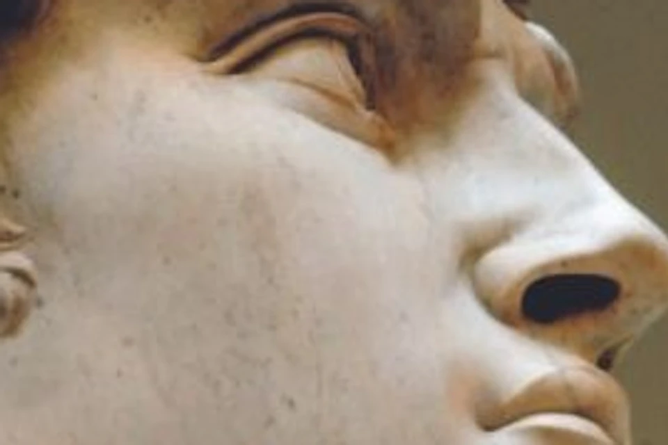 Close-up of a marble statue of a man's face with curly hair, holding an object near his chest.
