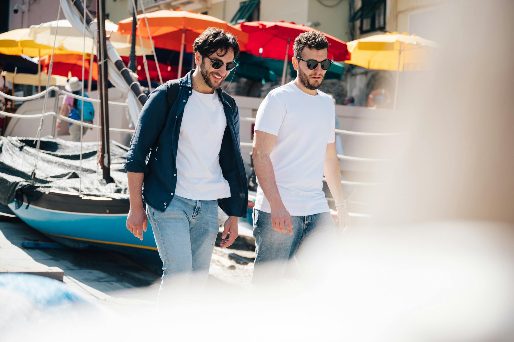 两名身着休闲服装和太阳镜的男士在户外散步，靠近五颜六色的雨伞和建筑物。