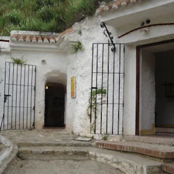 Vitväggig grottentré med järnportar och en stenväg, omgiven av grönska och rustika strukturer.