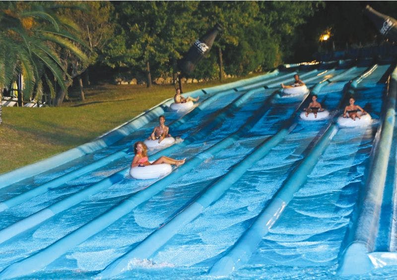 Three people in inflatable tubes sliding down a multi-lane water slide, surrounded by grass and trees.