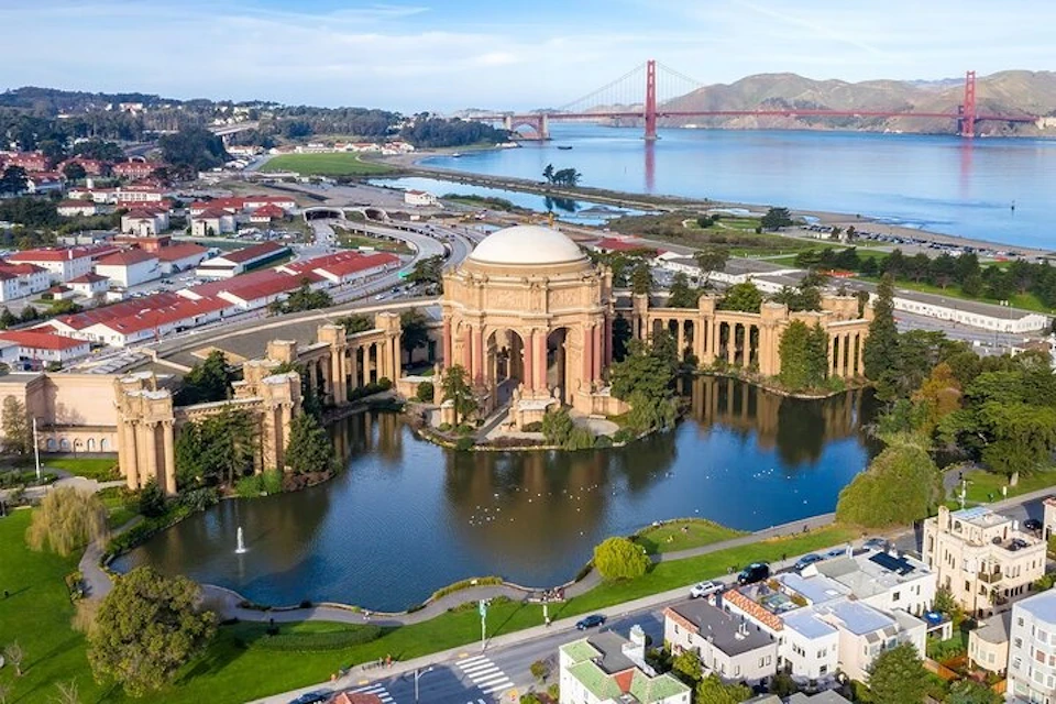 Aerial view of the Palace of Fine Arts in San Francisco, with a pond, nearby buildings, and the Golden Gate Bridge in the distance.