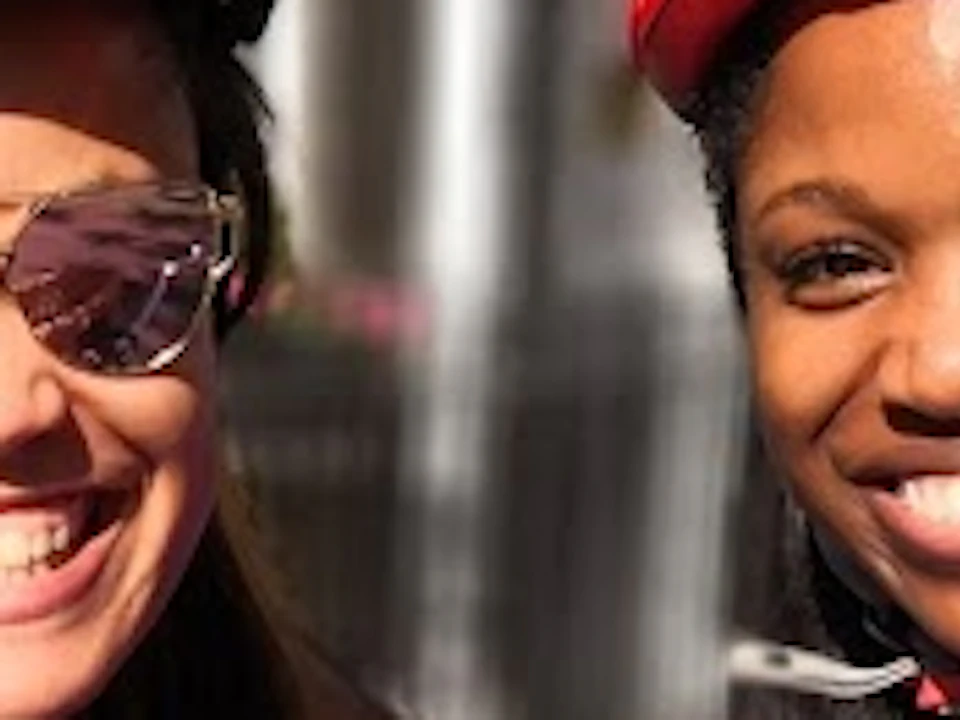 Two women, smiling, wearing helmets; one in a pink tank top, the other in a purple jacket, beside a bike with a wicker basket.