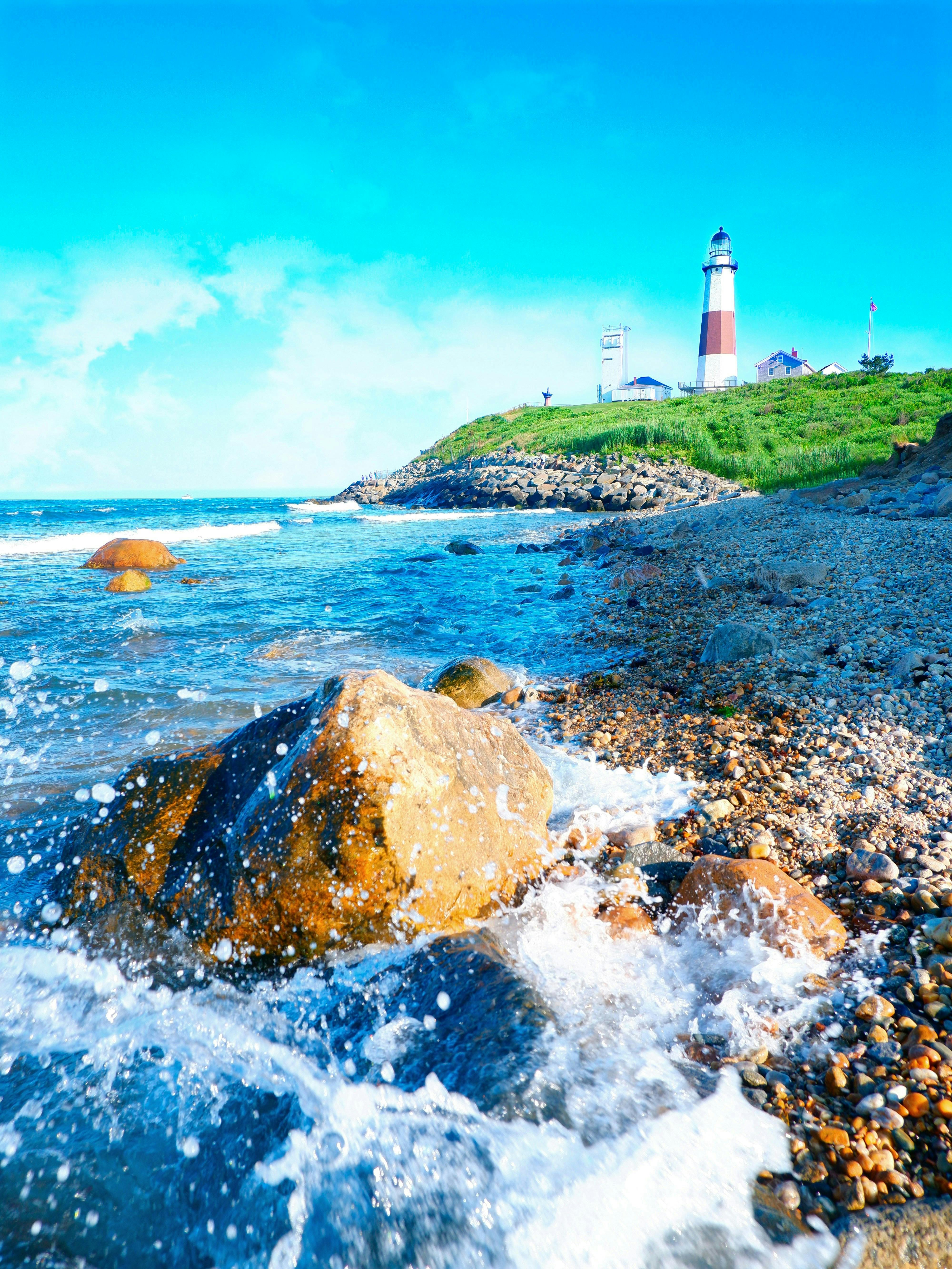 Waves crashing on a rocky shoreline with a lighthouse and buildings on a grassy hill under a bright blue sky.