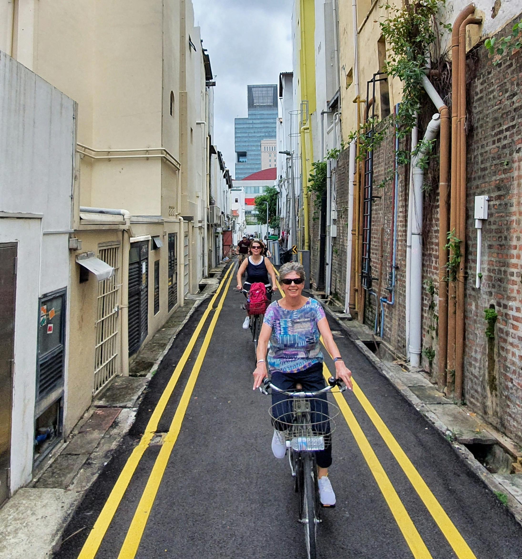 To personer cykler ned ad en smal gyde med bygninger, rør og gule vejmarkeringer. En høj bygning er synlig i det fjerne.