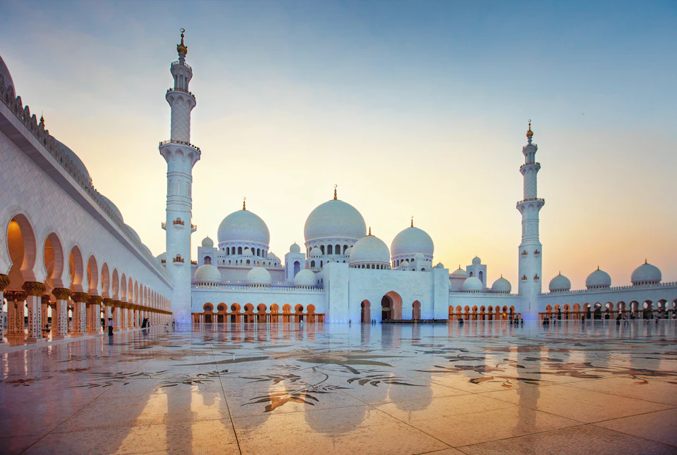 Grand mosque with multiple white domes and tall minarets at sunset, featuring intricate floral patterns on the courtyard floor.