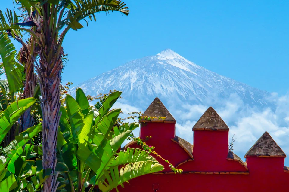 Snow-capped mountain behind a red building with pointed roofs, framed by green tropical foliage under a clear blue sky.