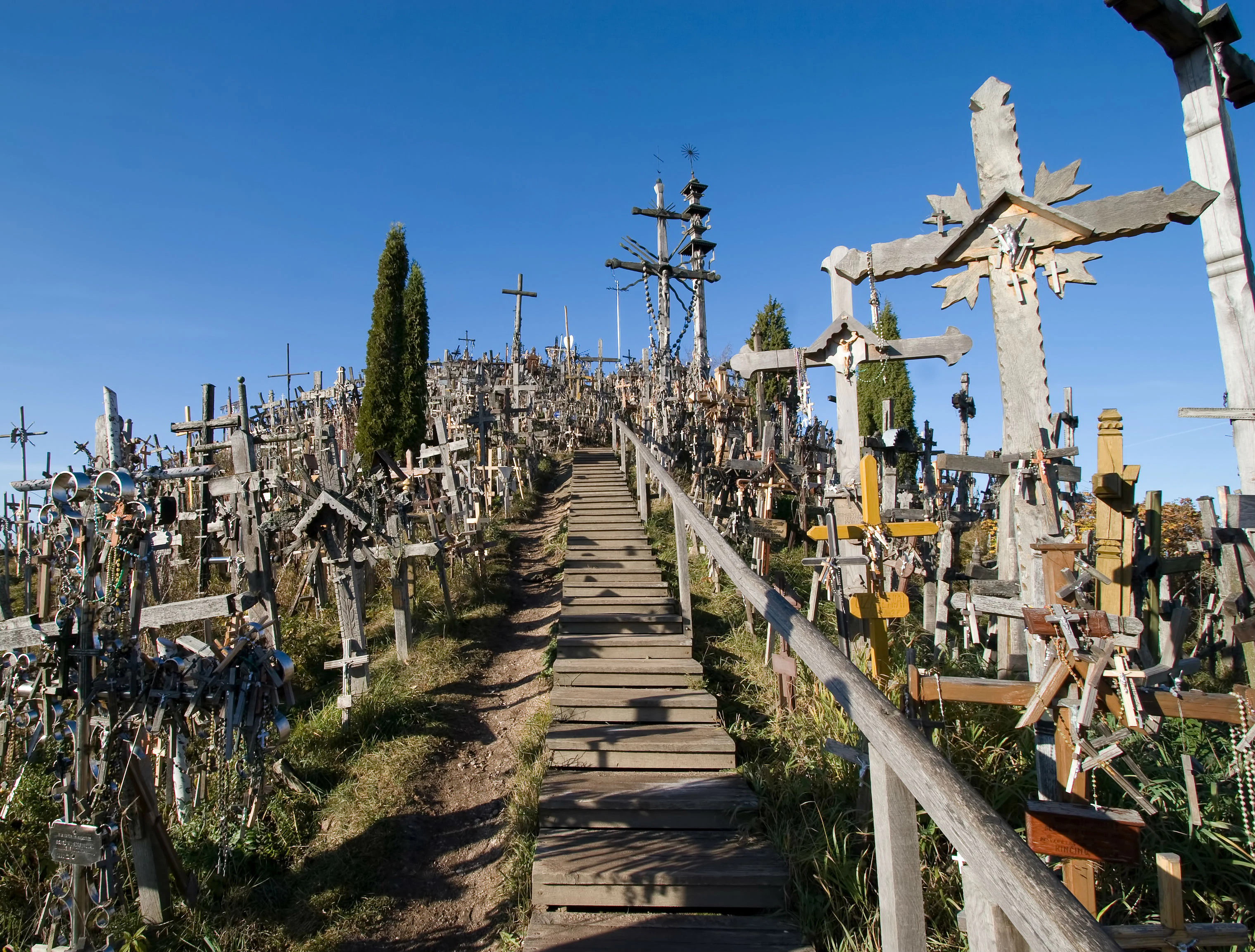 A hill covered with numerous crosses and a wooden path leading to the top under a clear blue sky.