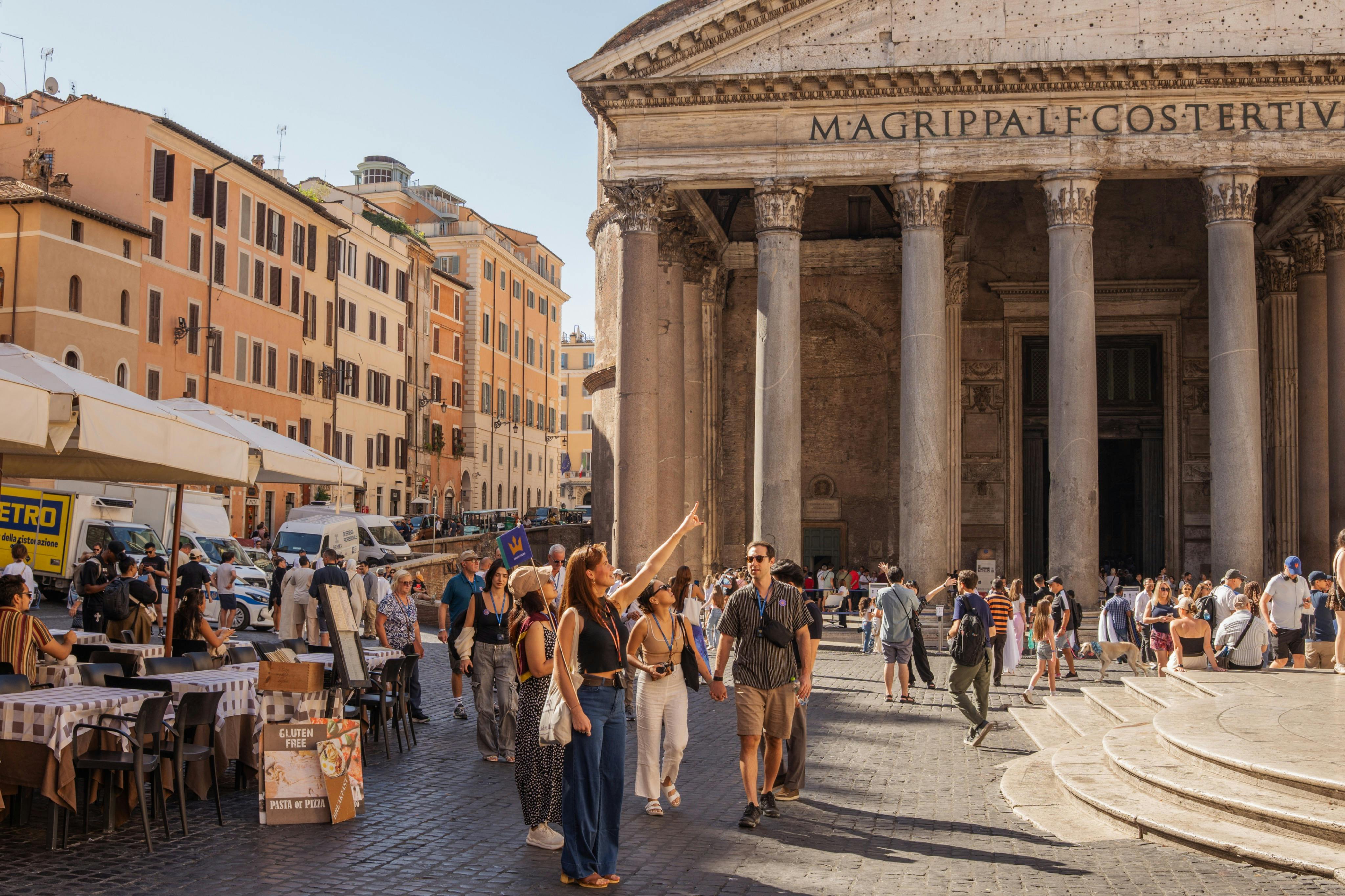 I turisti passeggiano e scattano foto davanti al Pantheon, circondati da edifici storici e caffè all&#39;aperto.