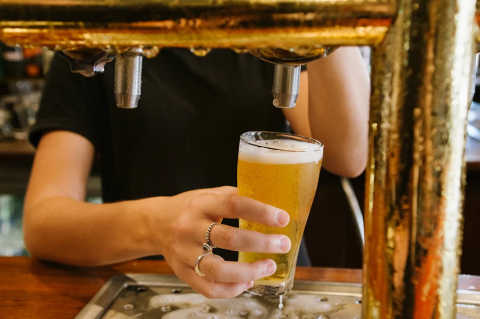A person pouring a glass of beer from a tap, holding the glass at an angle under a golden dispenser.
