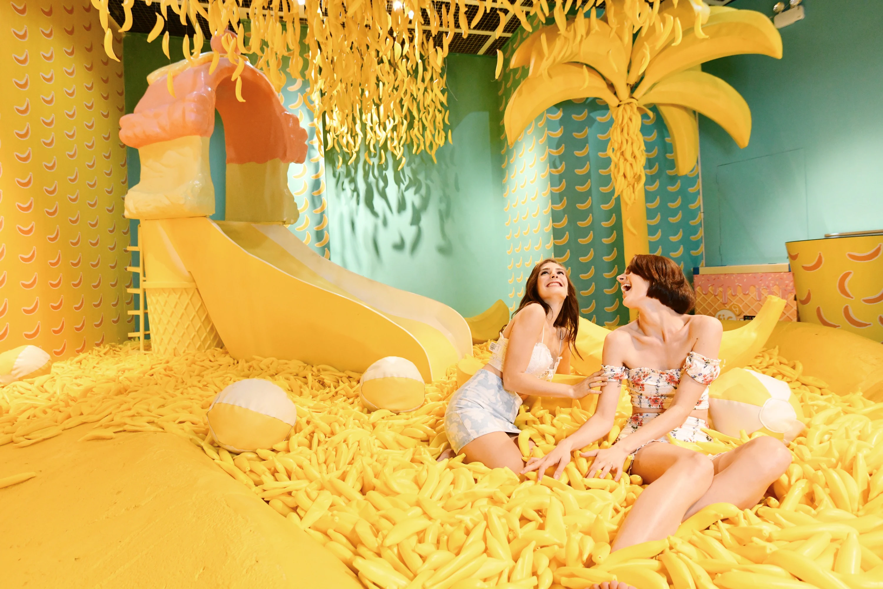 Two women laughing while sitting in a room filled with yellow banana-themed decor, including a banana tree and a slide.