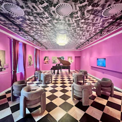 Ett rum med rosa tema med porträtt på väggarna, ett flygel i mitten, moderna stolar, rutigt golv och en ljuskrona.