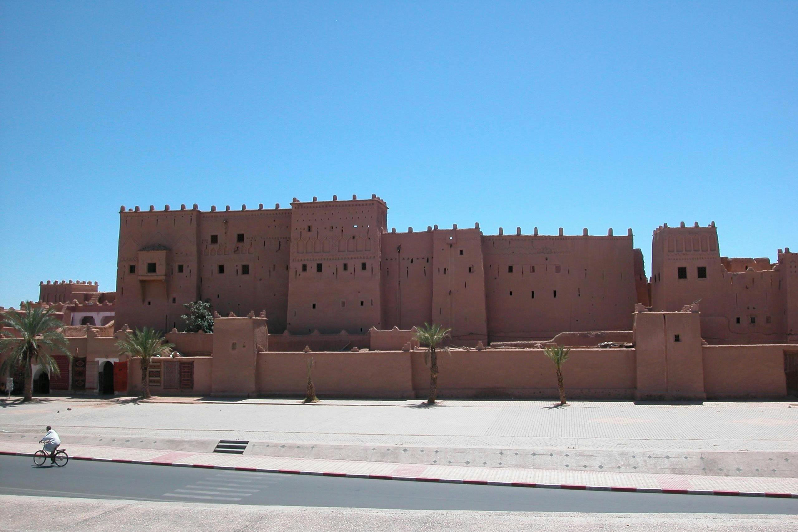 Une grande forteresse brune en briques de terre avec de petites fenêtres et des palmiers devant, sous un ciel bleu clair.