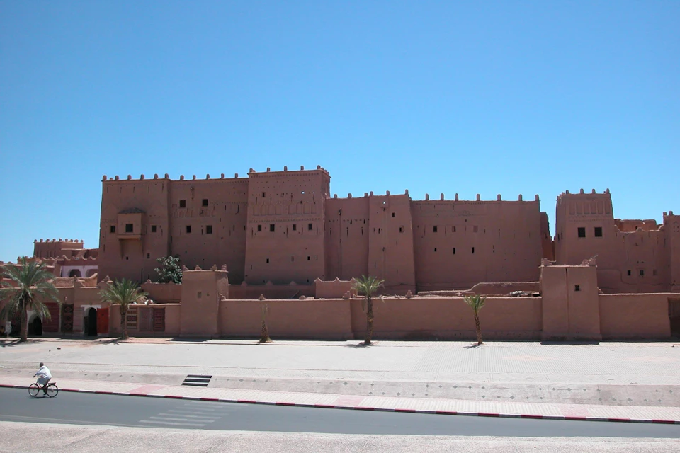 A large, brown, mud-brick fortress with small windows and palm trees in front, under a clear blue sky.