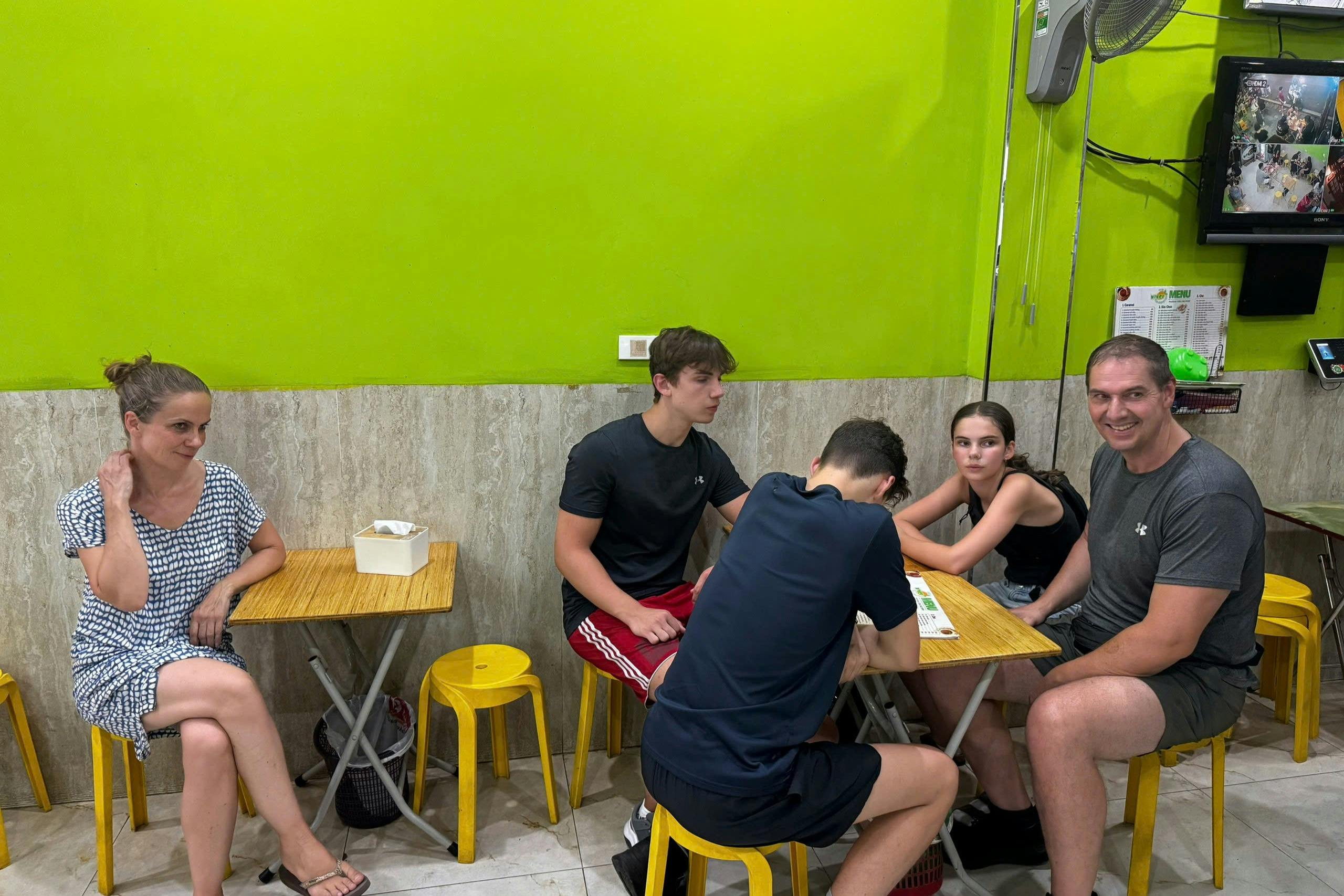 Four people sitting at a small table with yellow stools in a brightly lit room with lime green walls and a wall-mounted TV.