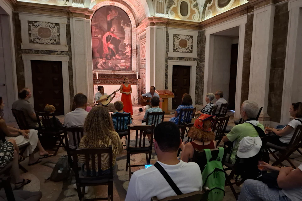 A small musical performance with three musicians and an audience seated in an ornate, historic room with a mural backdrop.