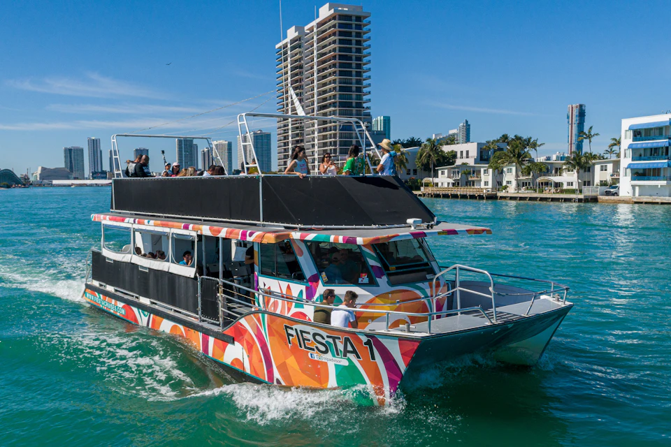 A colorful boat named "Fiesta 1" on turquoise water with people on board, city buildings and palm trees in the background.