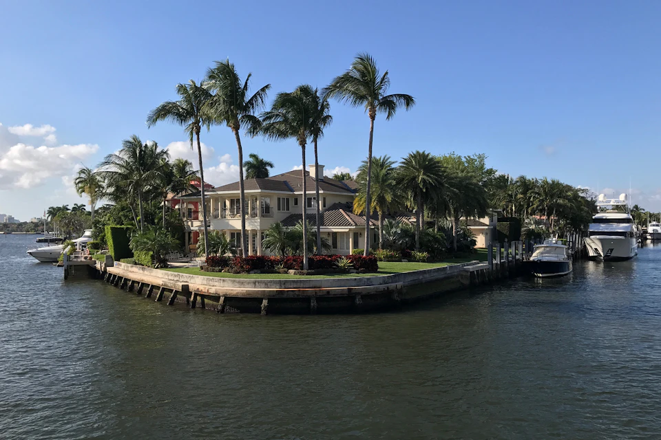 A waterfront house with palm trees, lush gardens, and a boat dock along a sunny shoreline.