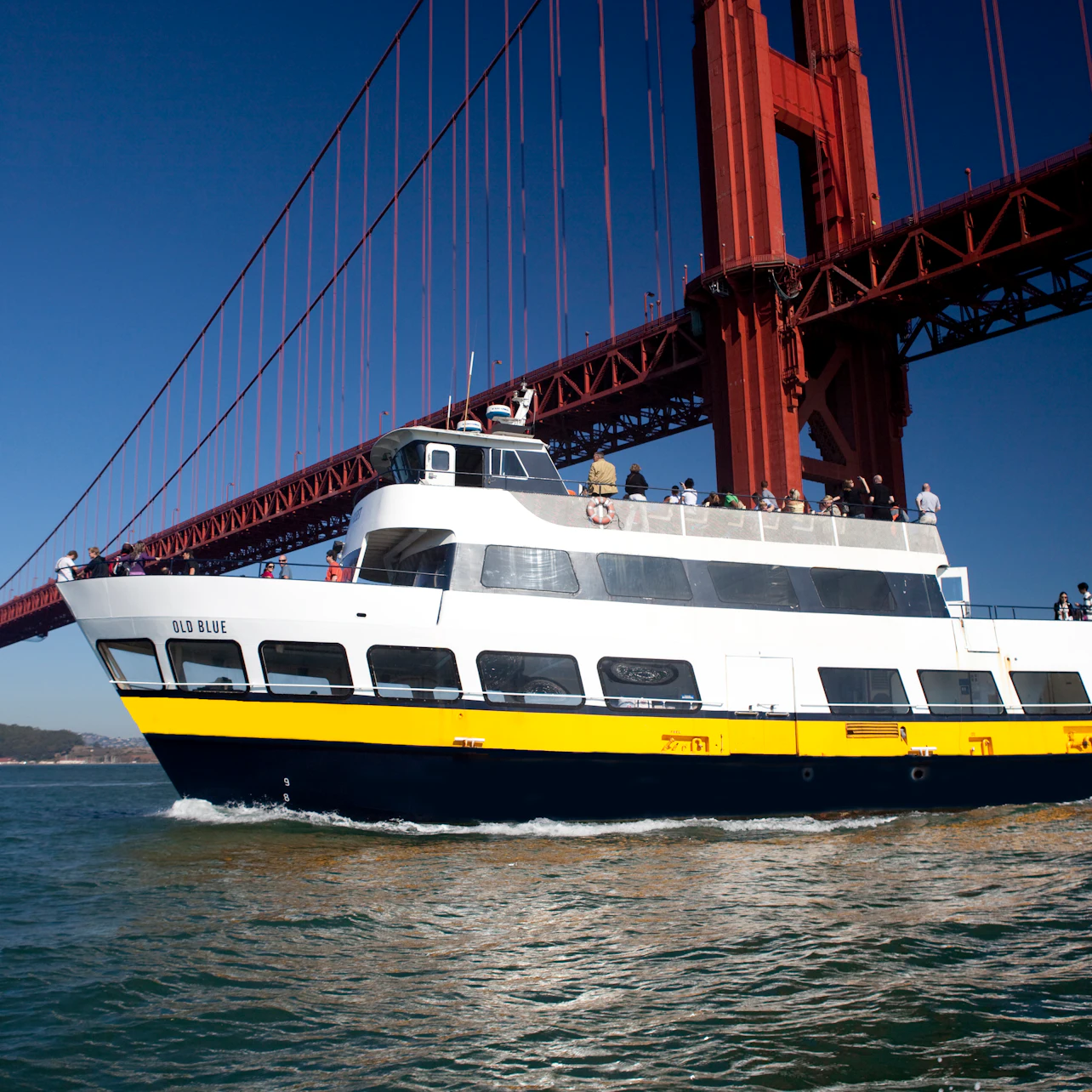 A white and yellow boat with passengers passes under the Golden Gate Bridge on a clear day.