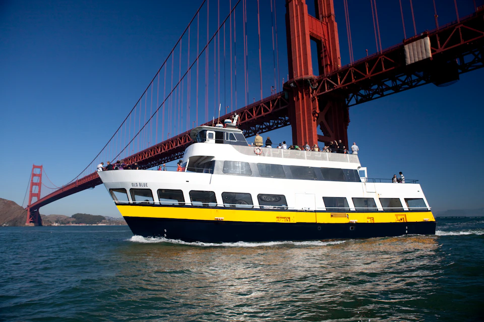 A white and yellow boat with passengers passes under the Golden Gate Bridge on a clear day.