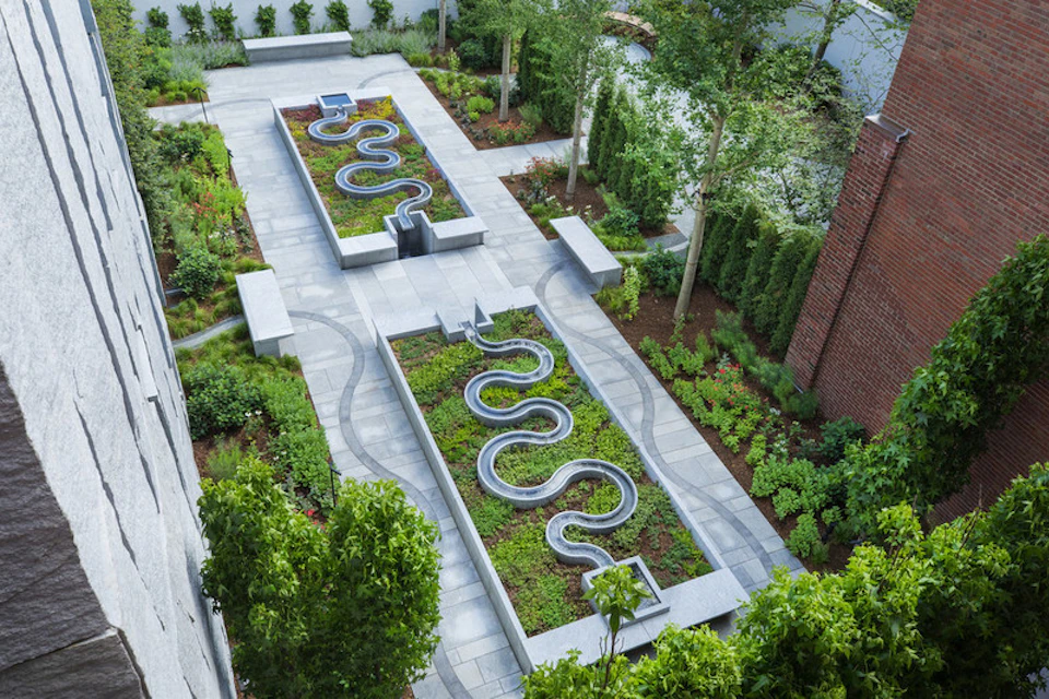 Modern garden with two long raised beds featuring curving metal water channels, surrounded by neatly landscaped greenery and paths.