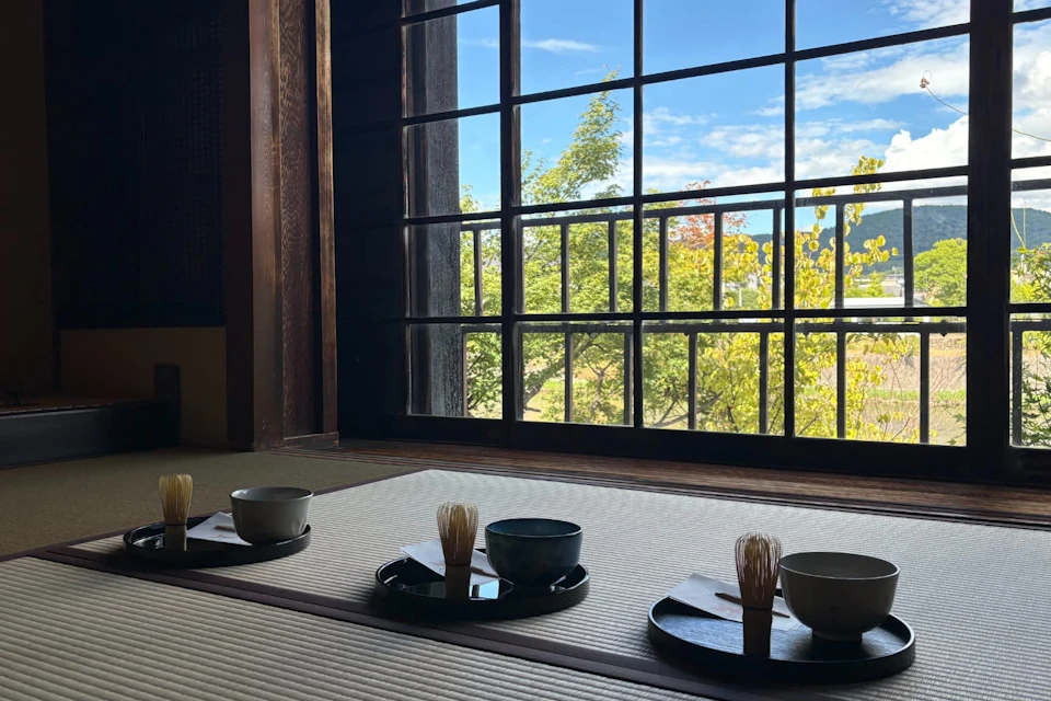 A traditional Japanese room with tatami floors, tea bowls arranged on trays, and a large window overlooking a garden.
