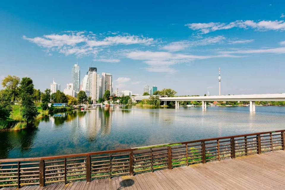 Cityscape with high-rise buildings, a wide river, a bridge, and a wooden viewing platform in the foreground under a clear blue sky.