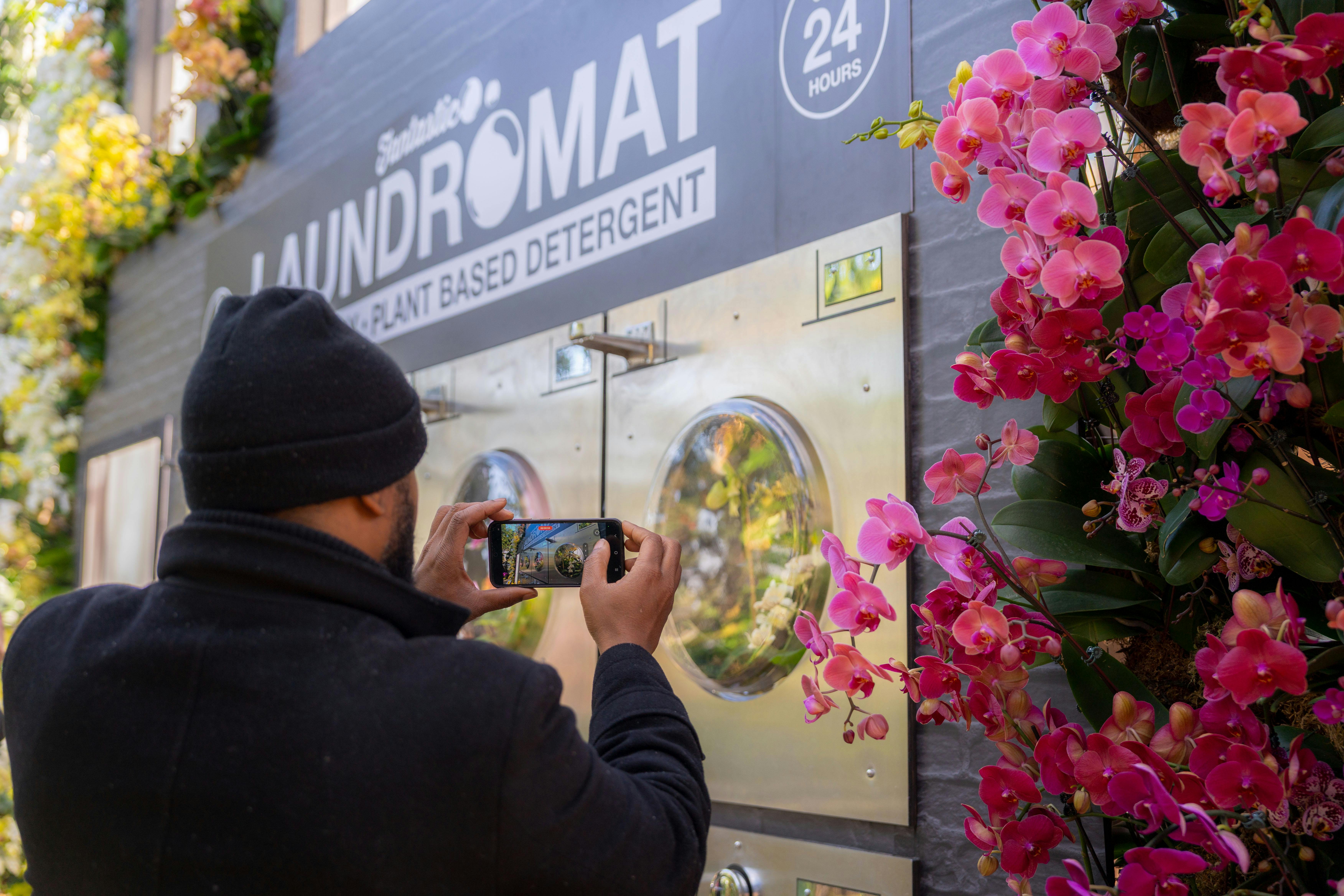 Una persona fotografiant una bugaderia amb un cartell que anuncia detergent a base de plantes i orquídies roses en primer pla.