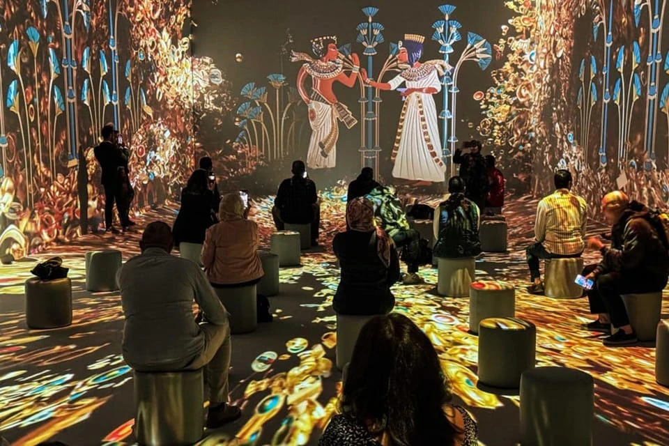 People sit on stools in a dimly lit room with colorful ancient Egyptian art projections on the walls and floor.