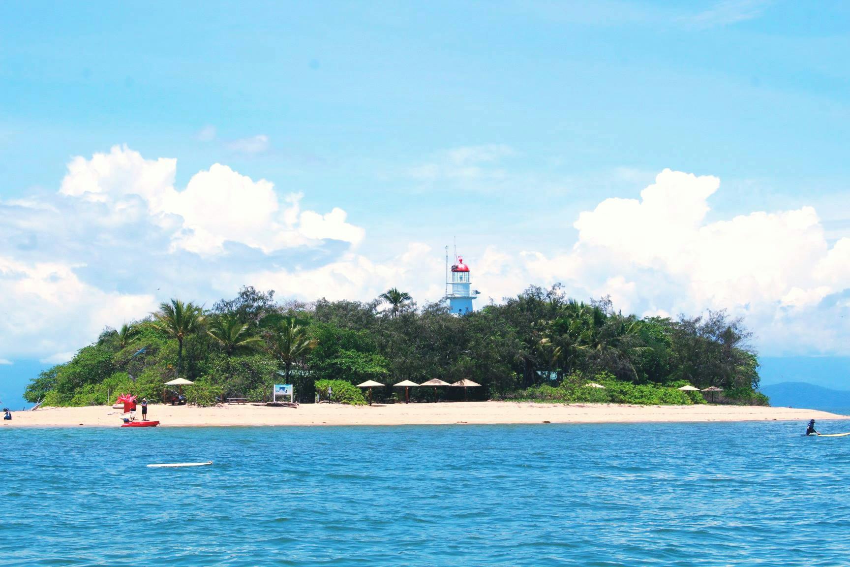 Sandy beach with palm trees, sun umbrellas, and a white lighthouse with a red roof in the background under a blue sky with clouds.
