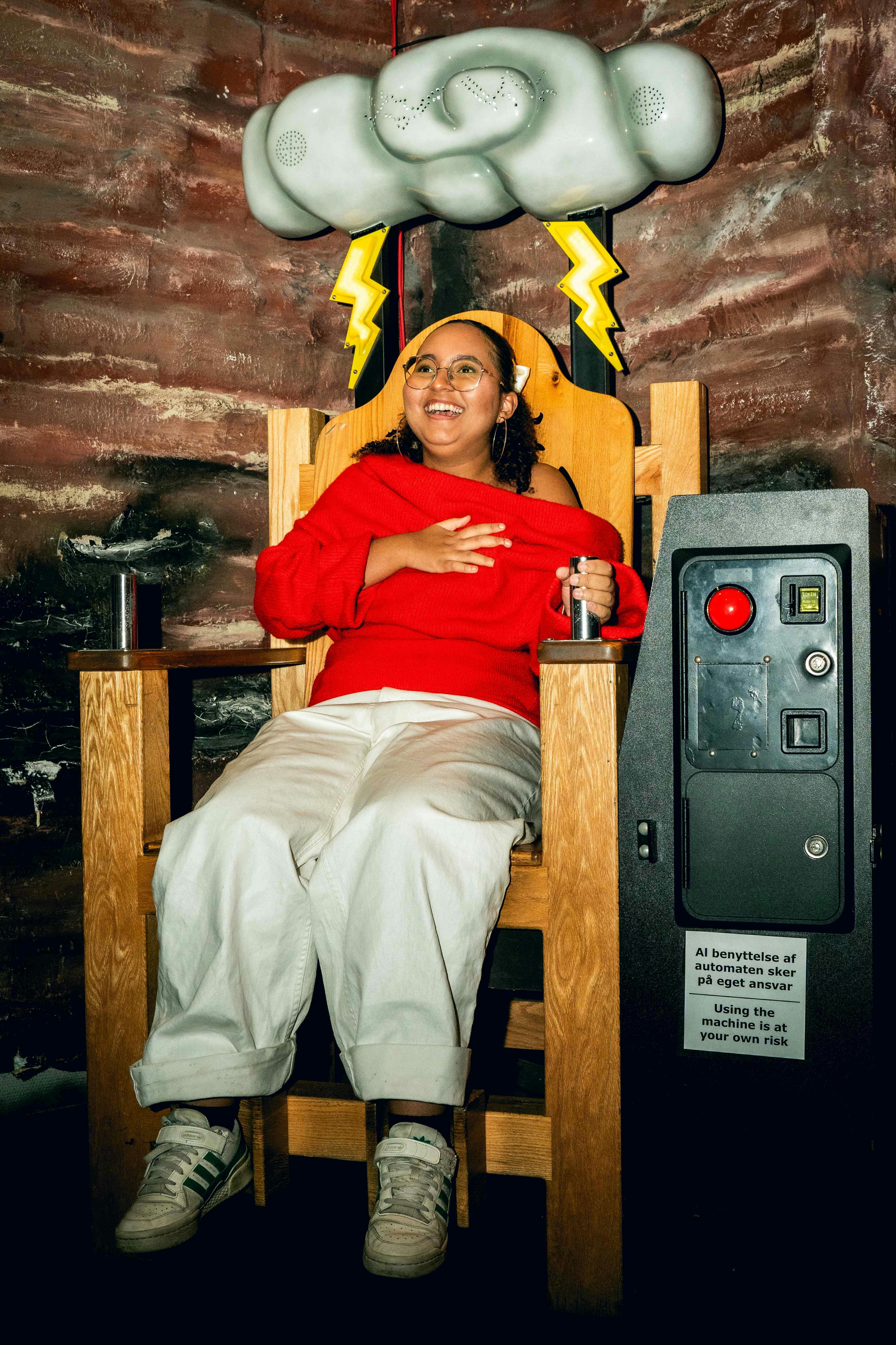 Person sitting in a wooden chair with lightning bolt decorations and a cloud structure overhead, smiling, and holding a lever.