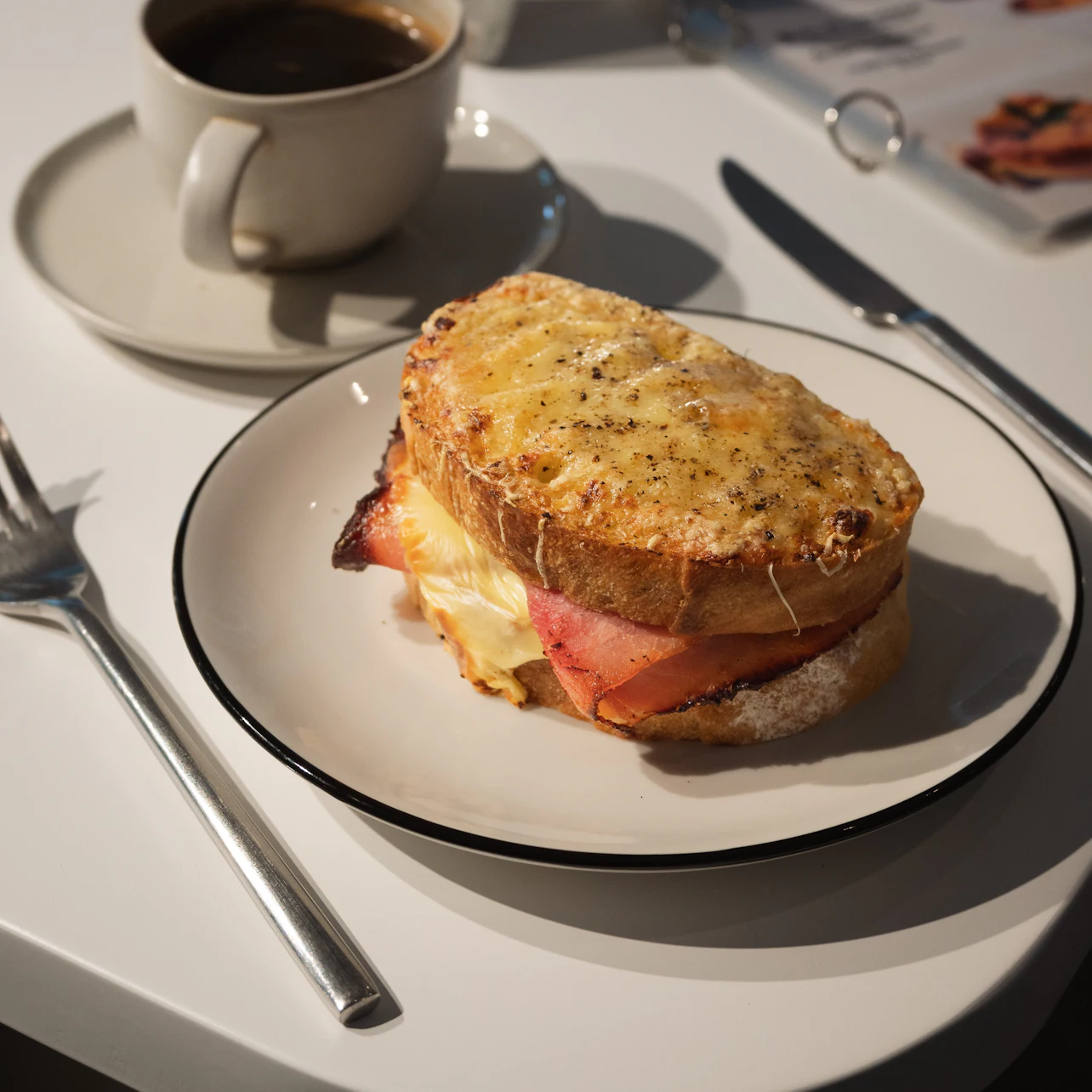 A grilled sandwich with ham and melted cheese on a plate, accompanied by a cup of coffee and utensils on a table.