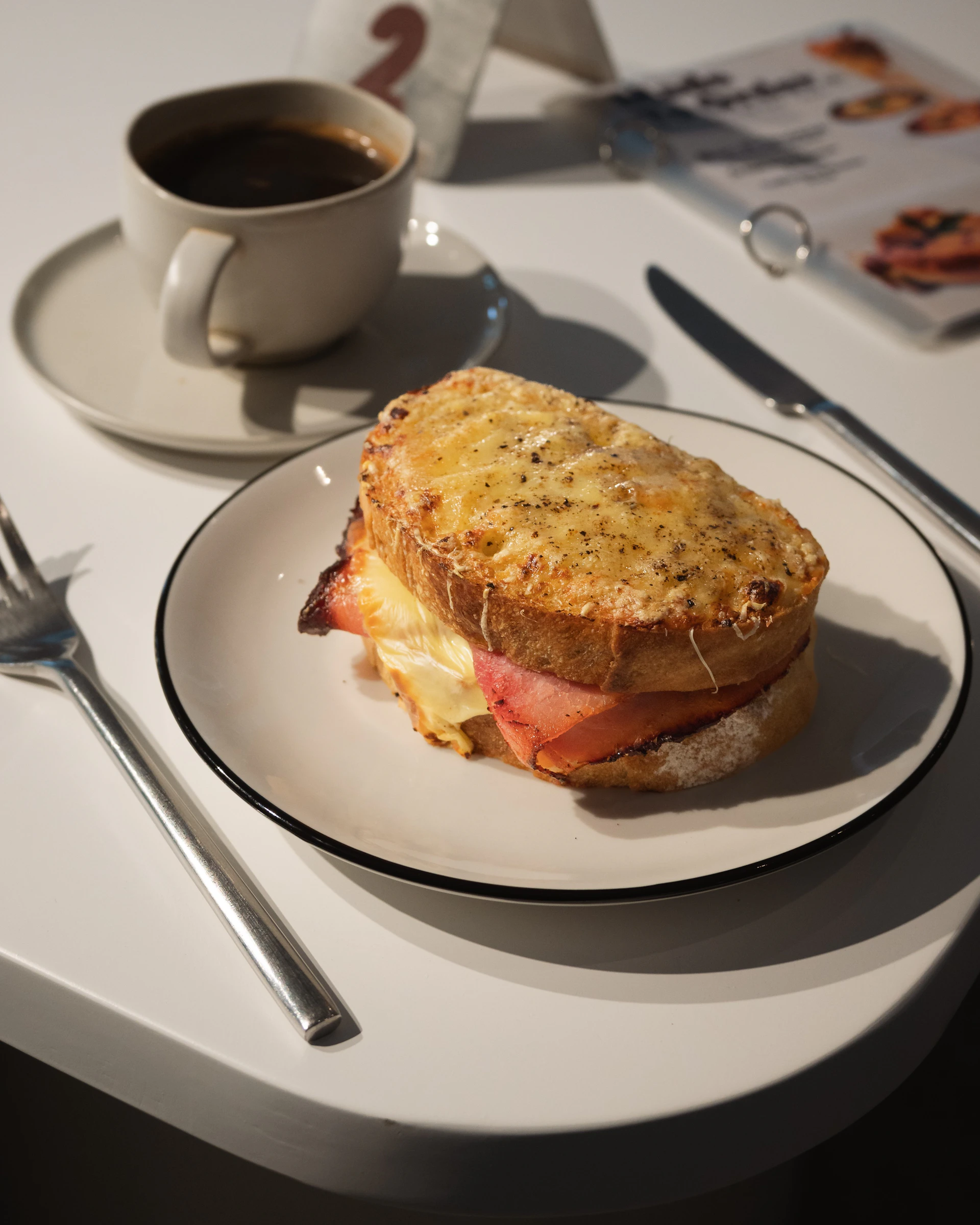 A grilled sandwich with ham and melted cheese on a plate, accompanied by a cup of coffee and utensils on a table.
