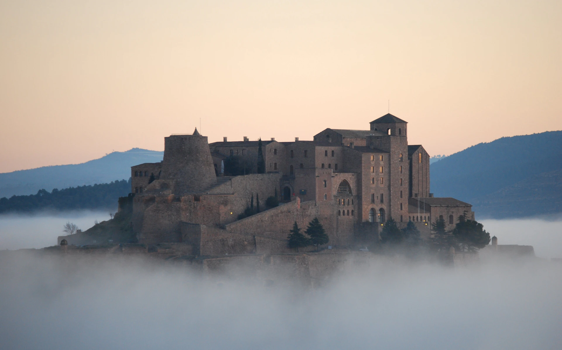 A large, historic stone castle with towers and arches stands atop a hill, emerging through thick fog with mountains in the background at dawn.