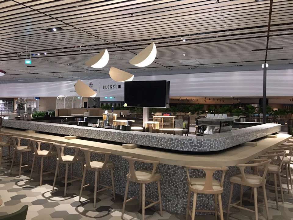 A modern bar with wooden stools, overhead crescent-shaped lights, and a central TV screen. The sign reads "Blossom."