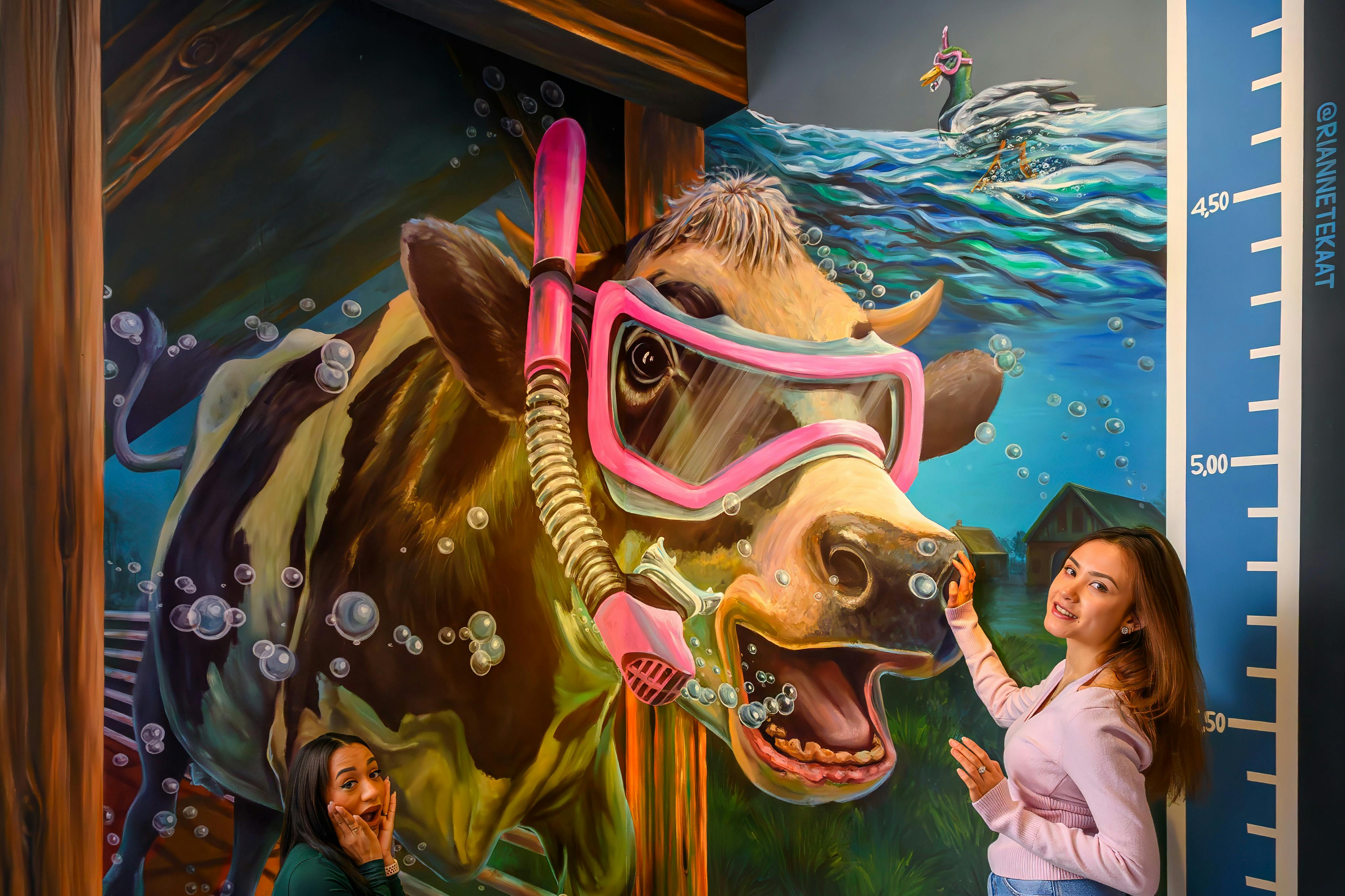Duas mulheres posam junto a um mural colorido de uma vaca com equipamento de mergulho e uma cena subaquática caprichosa como pano de fundo.