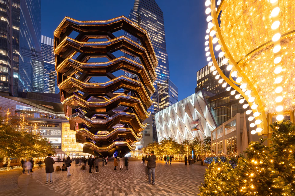 People walking near an illuminated, honeycomb-like structure and decorated tree in an urban plaza at dusk.